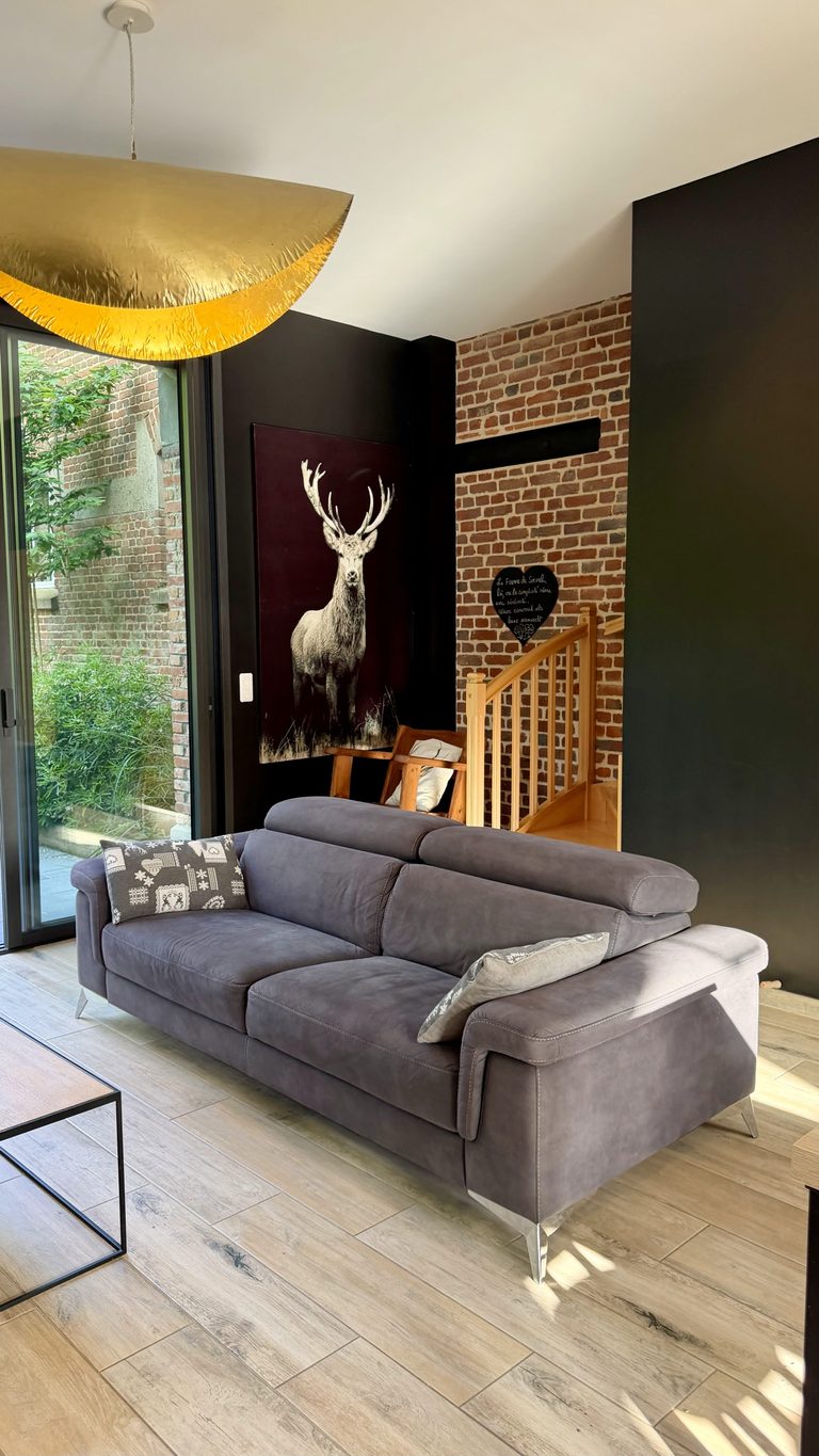 Salon avec canapé gris, mur en briques et tableau de cerf dans le cottage Aux Bois Dorés, Ferme de Sorval